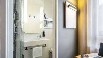 Een moderne badkamer met een glazen douchecabine, een witte wastafel en een spiegel erboven. Een handdoek hangt aan de muur en er staat een klein bureau met een laptop en zonnebril.