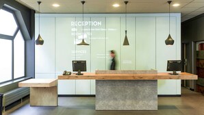 Un mostrador de recepción con encimera de madera, dos monitores de ordenador y un teléfono. Hay tres lámparas colgantes sobre el mostrador. El área de recepción tiene una pared de vidrio con texto que describe los servicios y tarifas de las habitaciones.
