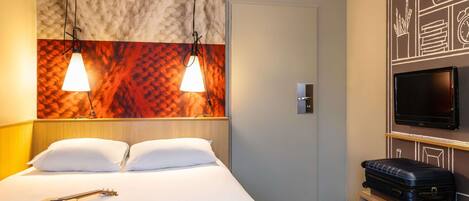 A hotel room with a bed, a wall-mounted TV, a suitcase, a guitar, and a magazine.