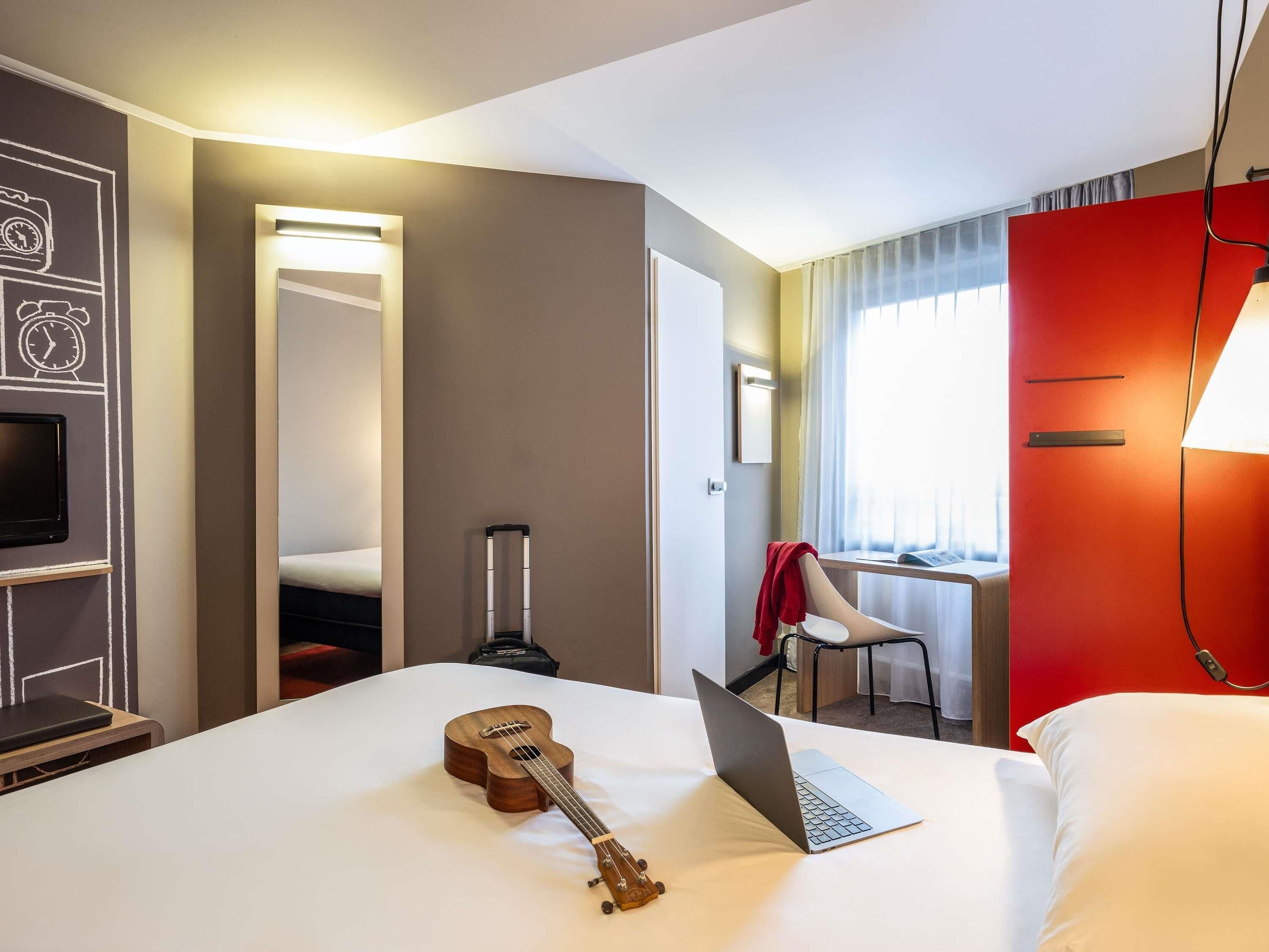 A hotel room with a bed, a laptop, a ukulele, a chair, and a red wall.