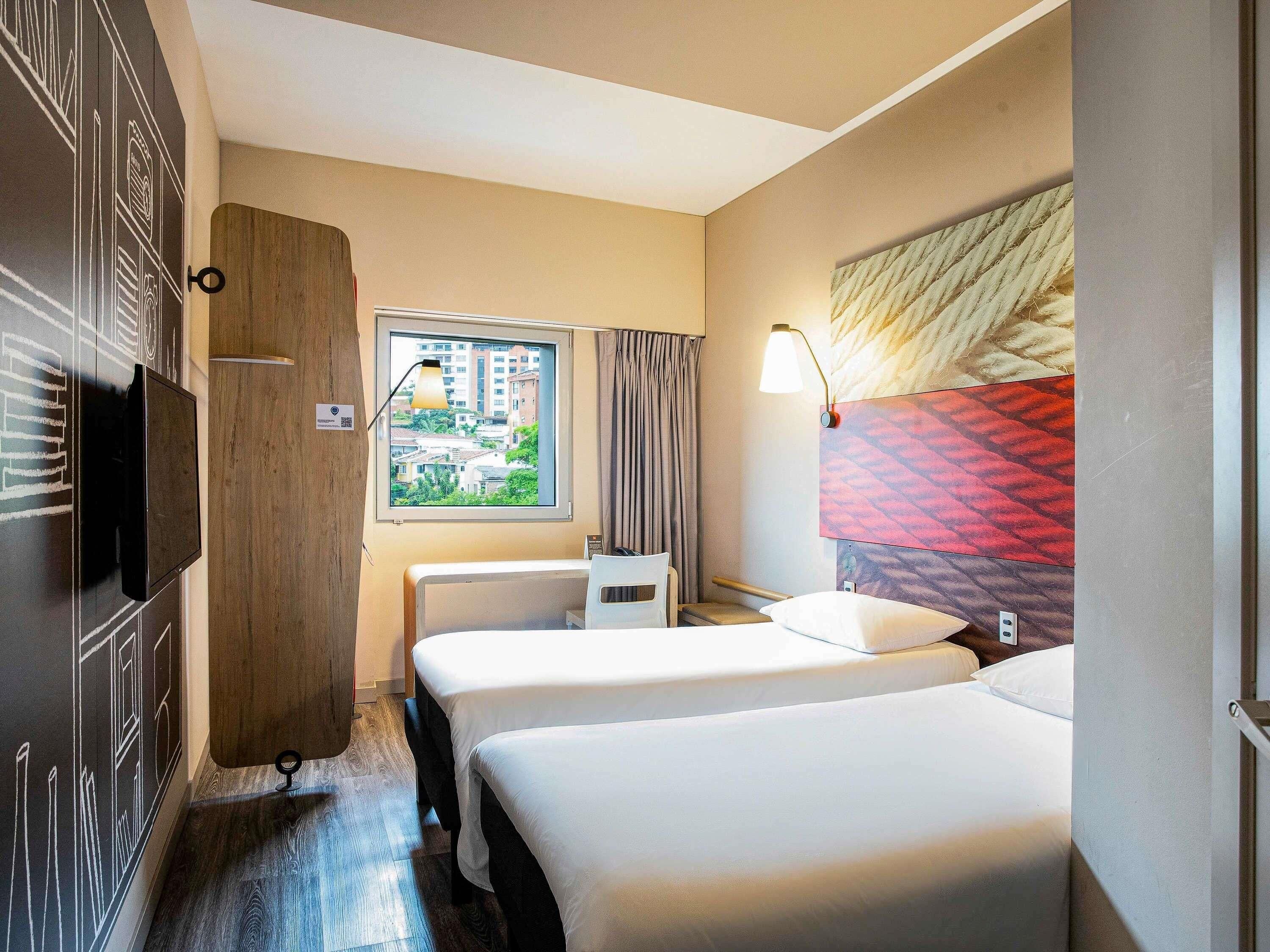 A hotel room with two beds, a TV, a window with a view of buildings, and a lamp.