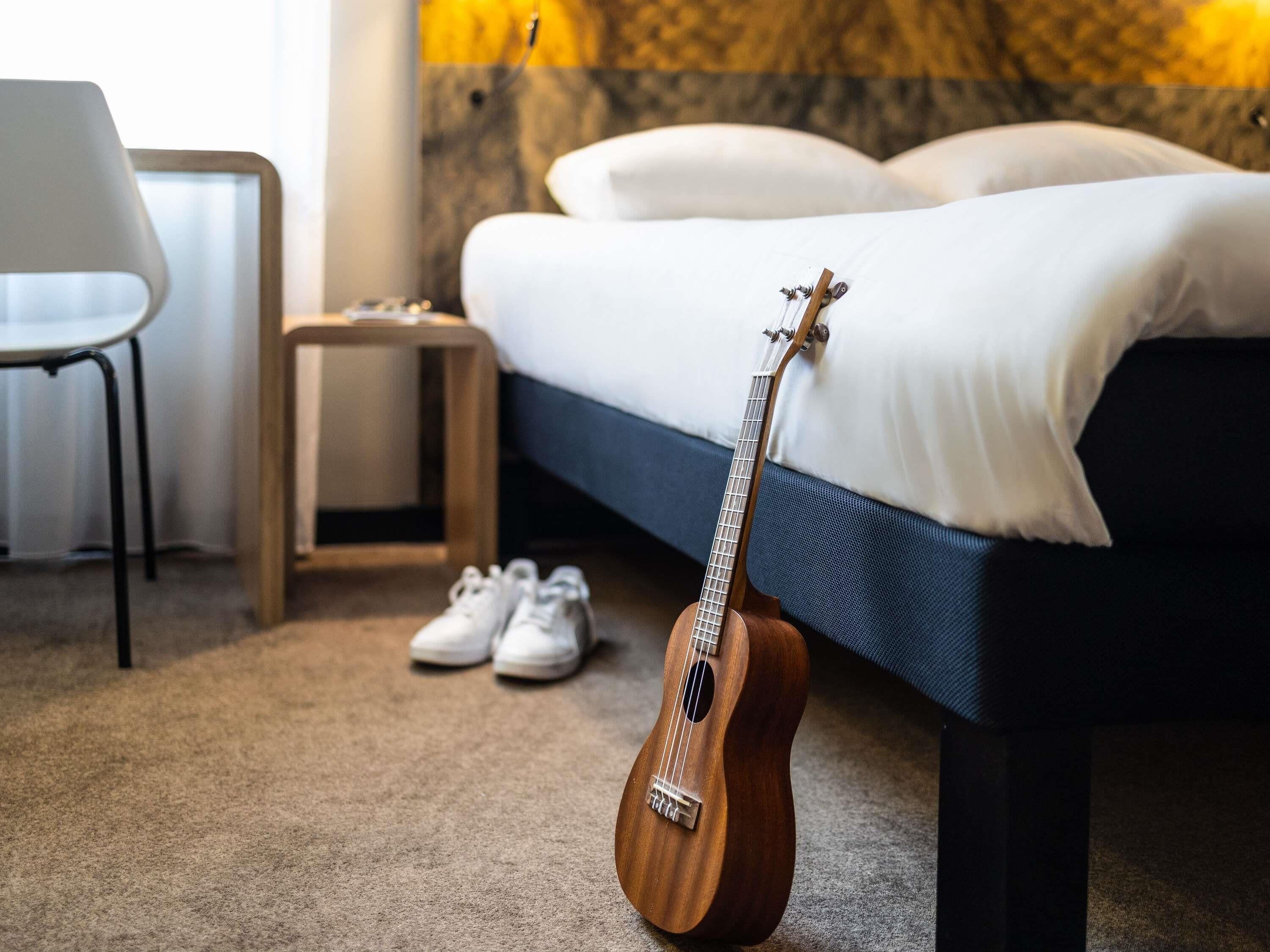 Una cama con un edredón blanco y almohadas, una guitarra de madera apoyada en ella, un par de zapatillas blancas en el suelo y una mesita de noche de madera con una lámpara.