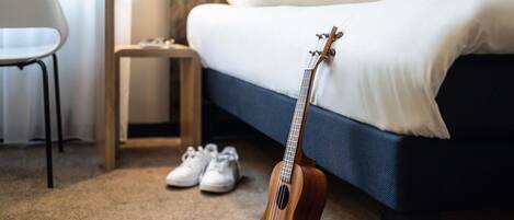 Una cama con un edredón blanco y almohadas, una guitarra de madera apoyada en ella, un par de zapatillas blancas en el suelo y una mesita de noche de madera con una lámpara.