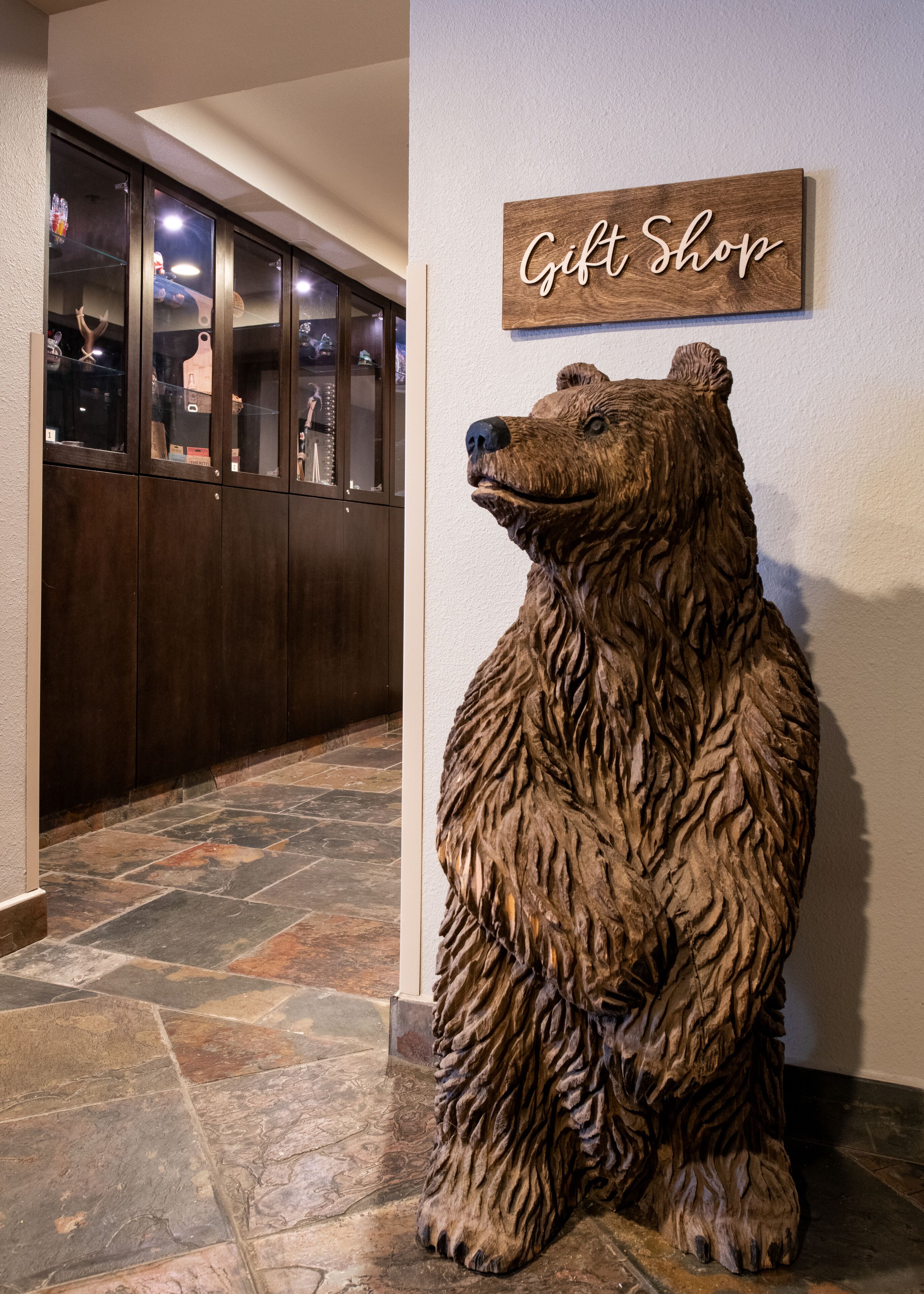 Un suelo de baldosas de piedra, un mueble de madera con puertas de cristal, y una escultura de madera de un oso con un letrero que dice 'Gift Shop'.
