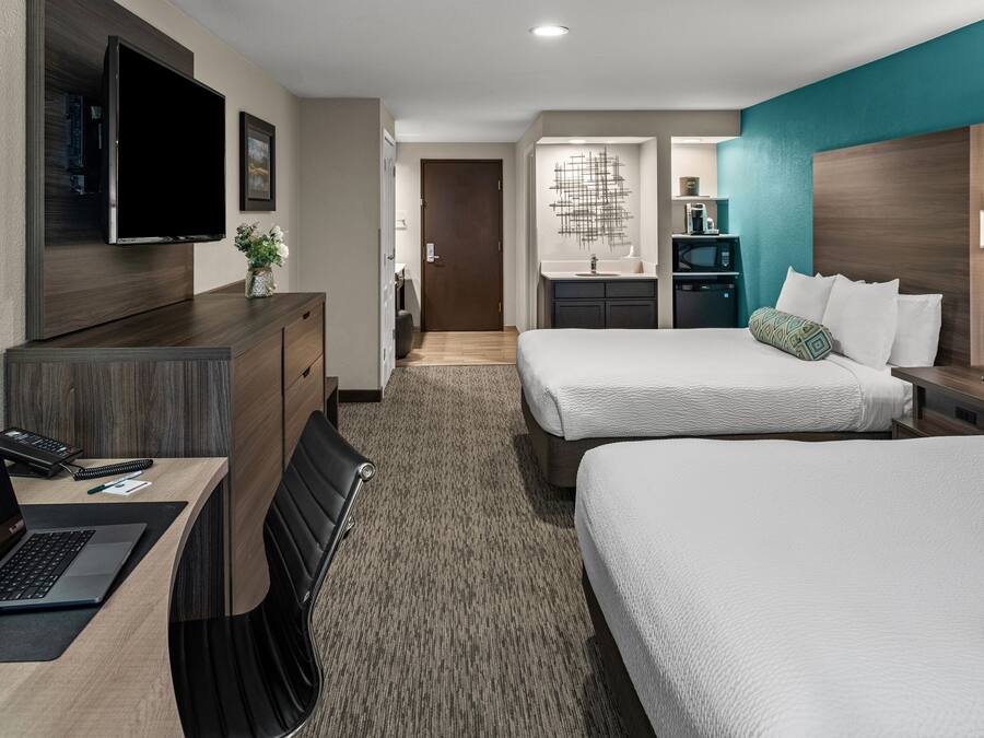 Habitación de hotel con dos camas, un escritorio con una computadora portátil, un televisor montado en la pared y un baño con lavamanos y espejo.