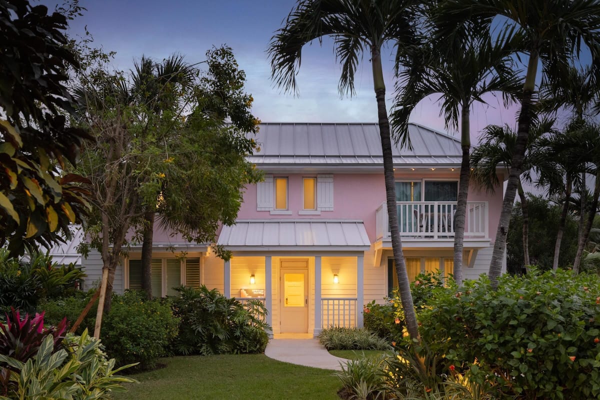 A pink house with a white balcony, surrounded by lush greenery and palm trees.