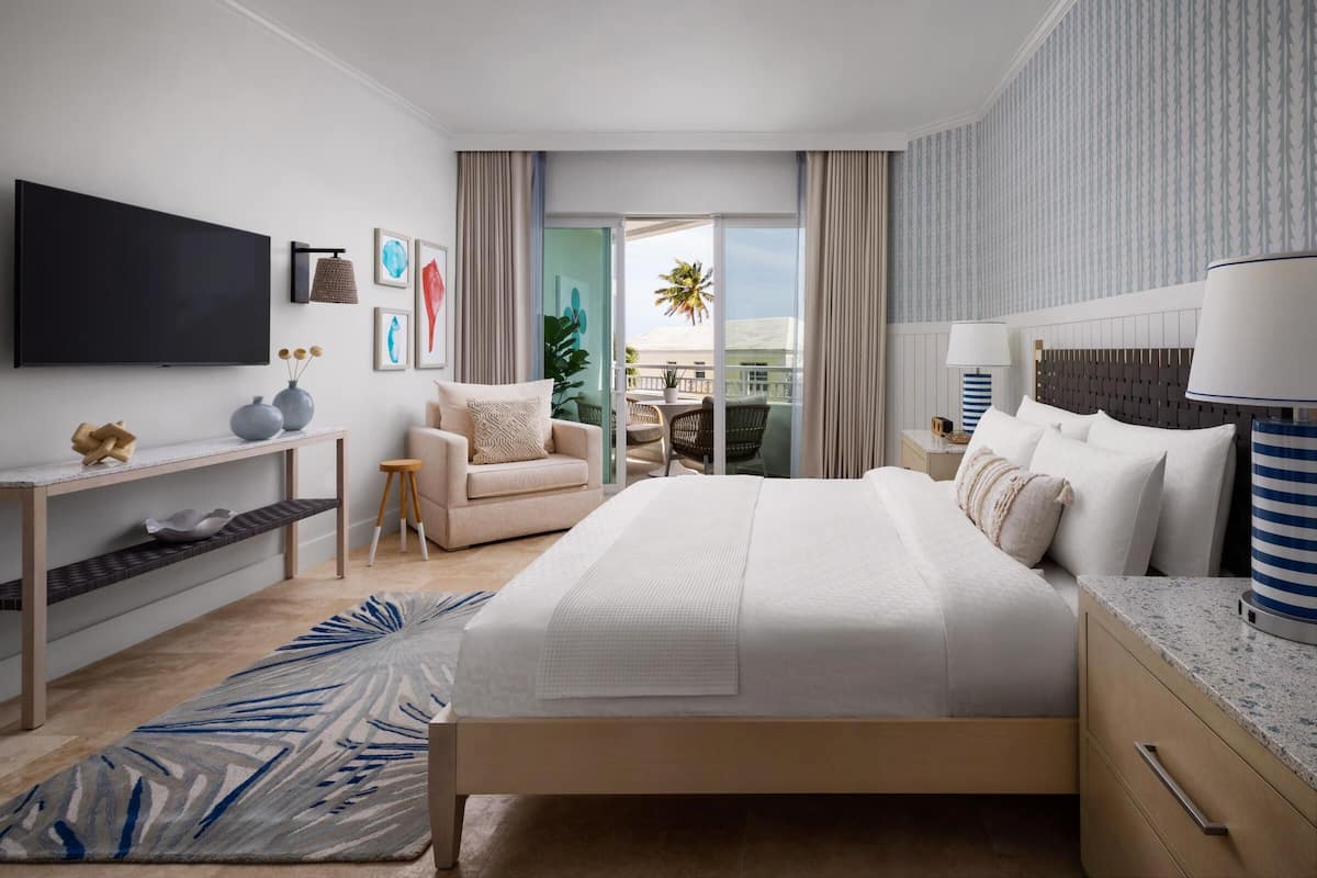 A modern hotel room with a large bed, a flat-screen TV, a balcony with a view of palm trees, and a dining area with wicker furniture.