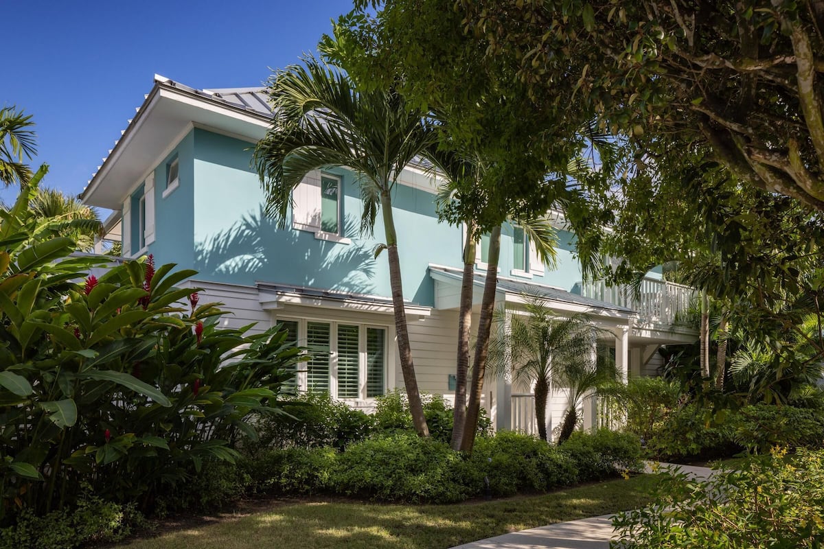 A two-story house with a light blue exterior, surrounded by lush greenery and palm trees.