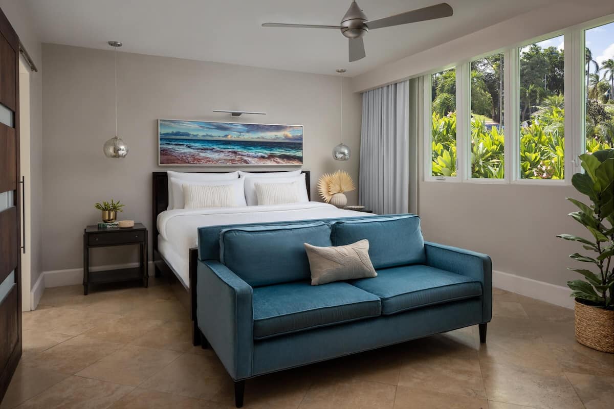 A bedroom with a bed, a blue sofa, a ceiling fan, a painting, and a window with a view of greenery.