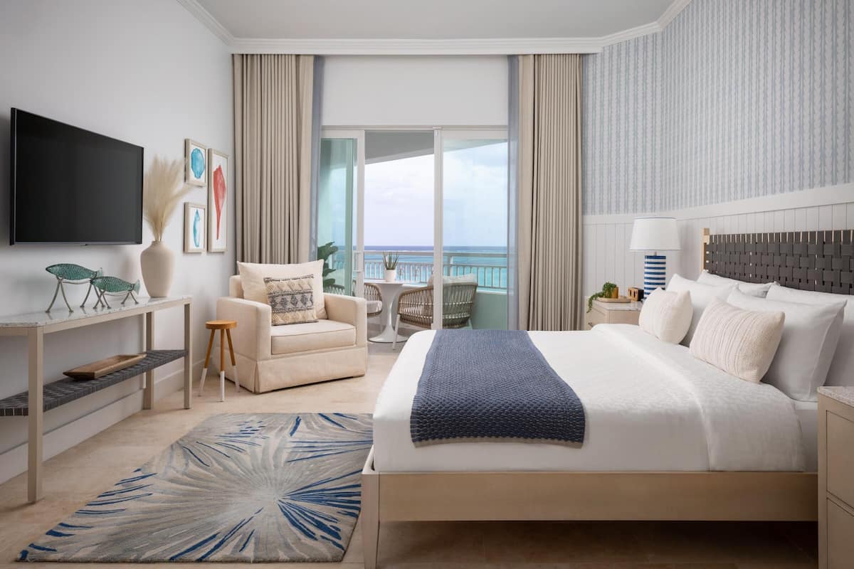 A hotel room with a large bed, a TV, a desk, a chair, a small table, and a view of the ocean.