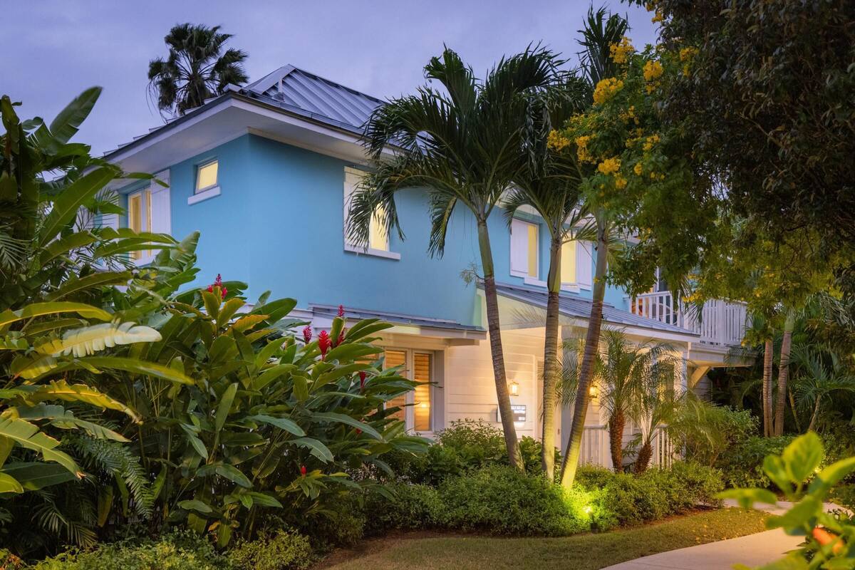 A two-story house with a blue exterior, surrounded by lush tropical vegetation, including palm trees and various plants.