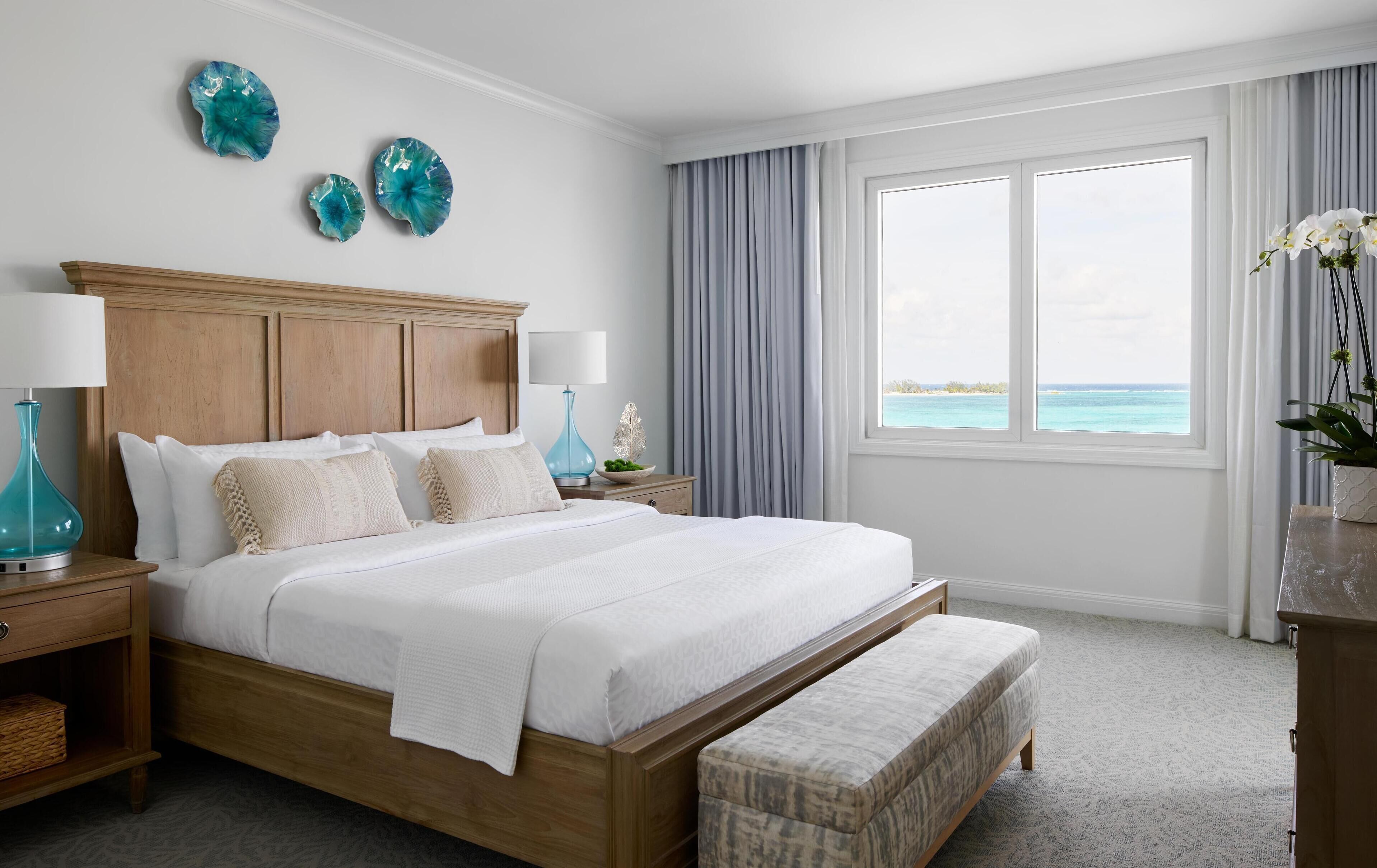 A spacious bedroom with a large bed, wooden headboard, two nightstands, a bench, and a view of the ocean through a large window.