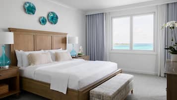 A spacious bedroom with a large bed, wooden headboard, two nightstands, a bench, and a view of the ocean through a large window.