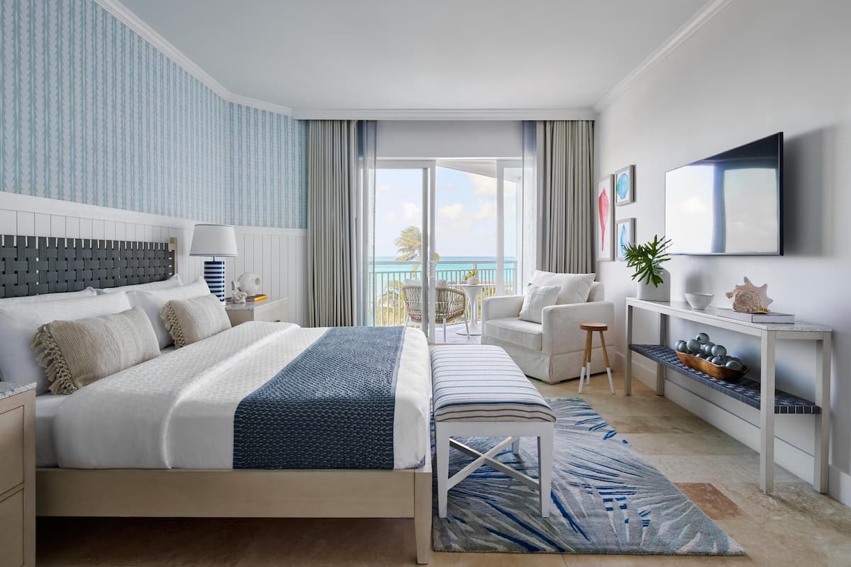 A modern hotel room with a large bed, a balcony with a view of the sea, a seating area with a chair and a small table, and a TV mounted on the wall.