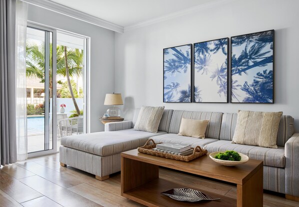 A modern living room with a sofa, a coffee table, and a view of a pool and palm trees through large windows.