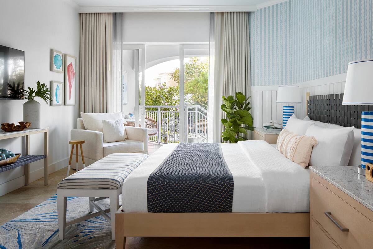 A modern hotel room with a large bed, a seating area with a chair and a small table, a balcony with a view, and a TV on the wall.