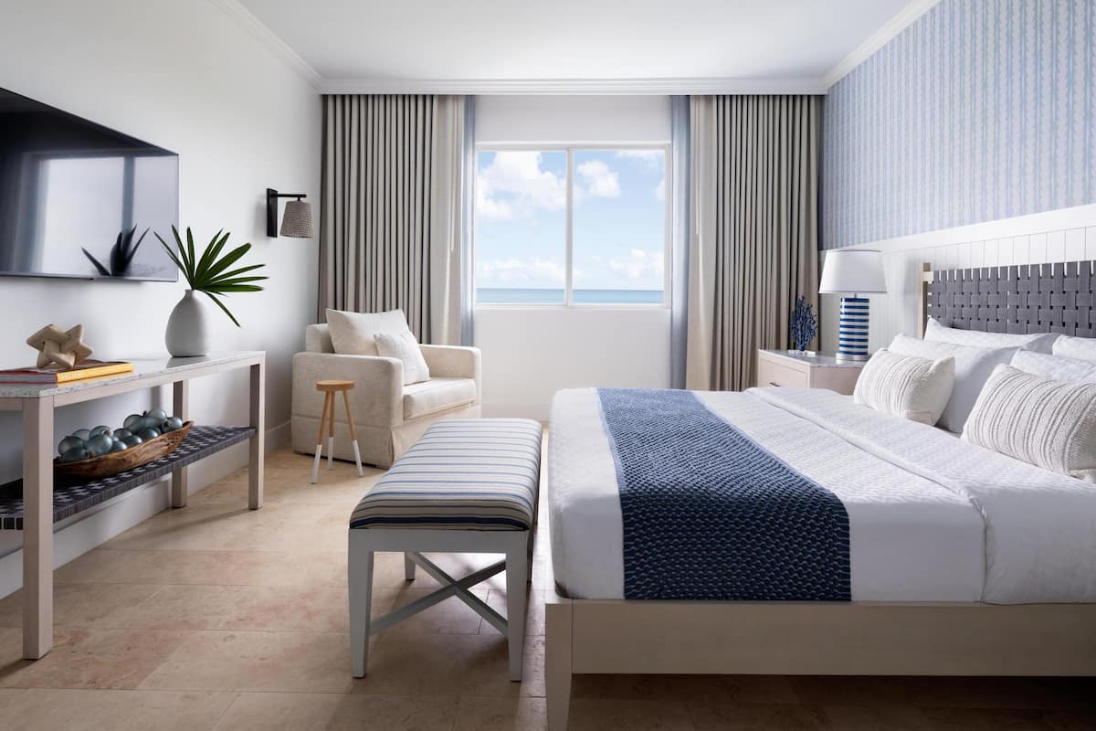 A modern hotel room with a large bed, a desk with a TV, a chair, and a view of the ocean.