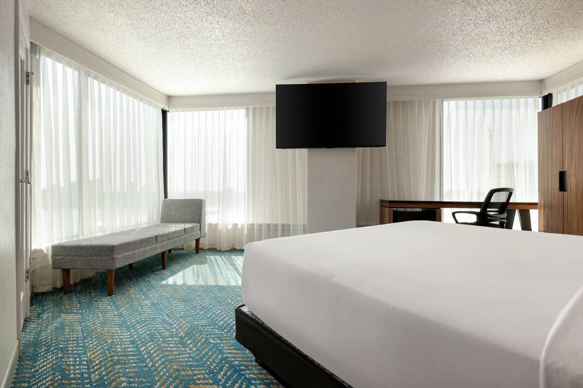 A hotel room with a large bed, a flat-screen TV mounted on the wall, a desk with a chair, and a bench by the window.