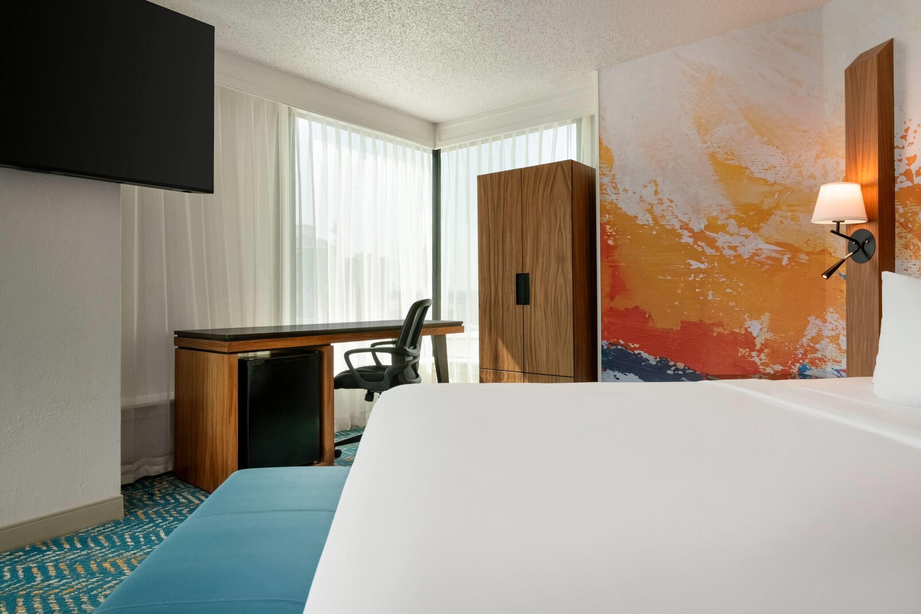 A hotel room with a bed, a desk, a chair, a TV mounted on the wall, and a large window with curtains.