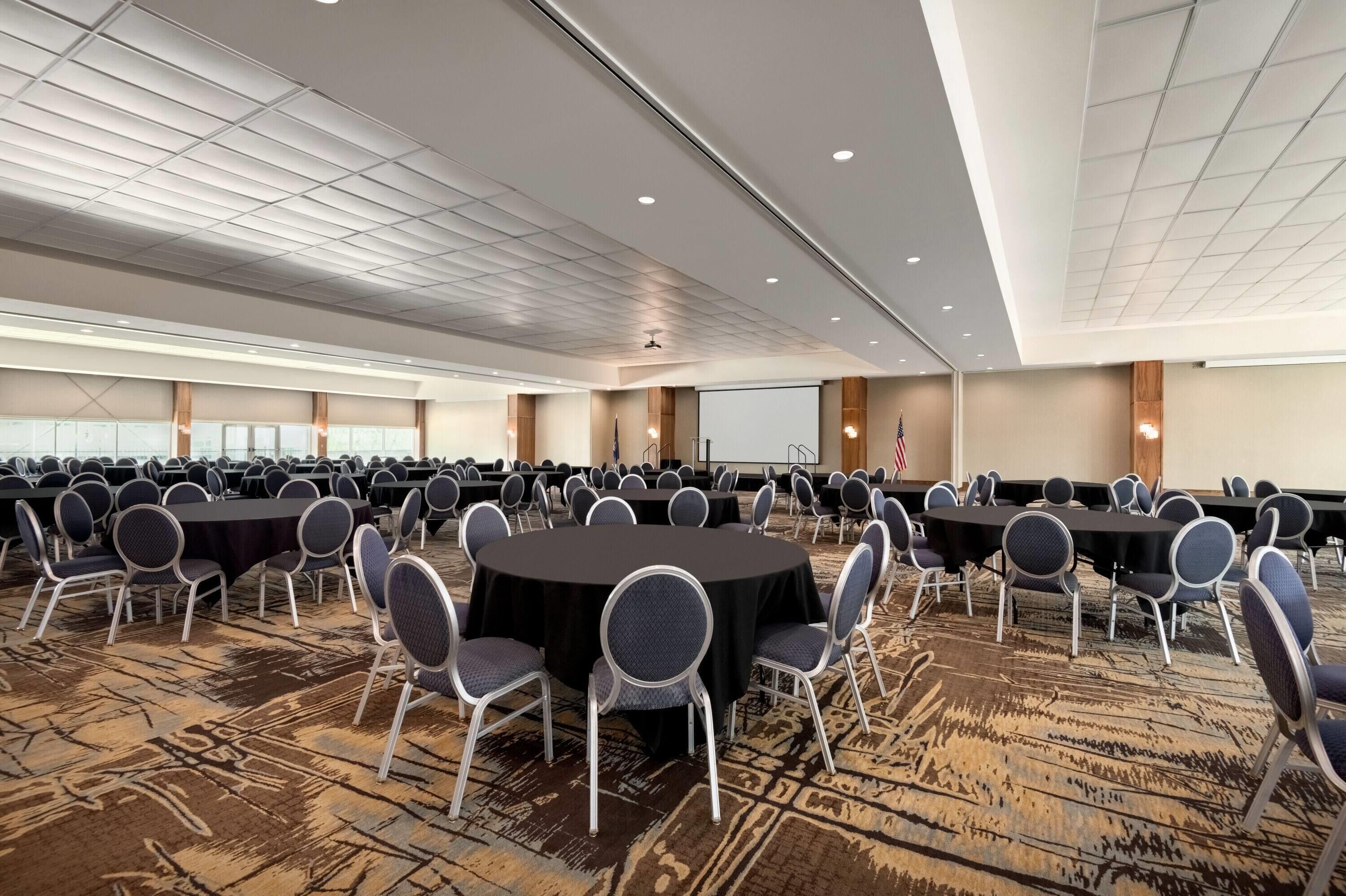 A large conference room with multiple round tables and chairs, a projector screen, and an American flag.