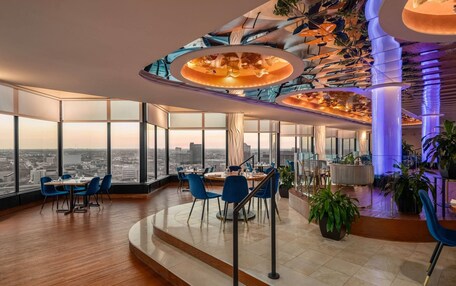 A modern hotel lobby with a high ceiling, large windows offering city views, a central seating area, and a staircase leading to an upper level.