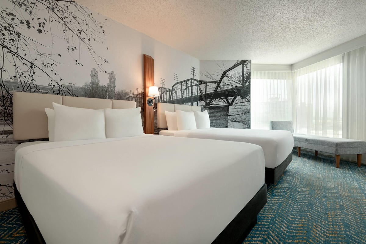 A hotel room with two beds, a large window with sheer curtains, and a wall mural of a bridge.