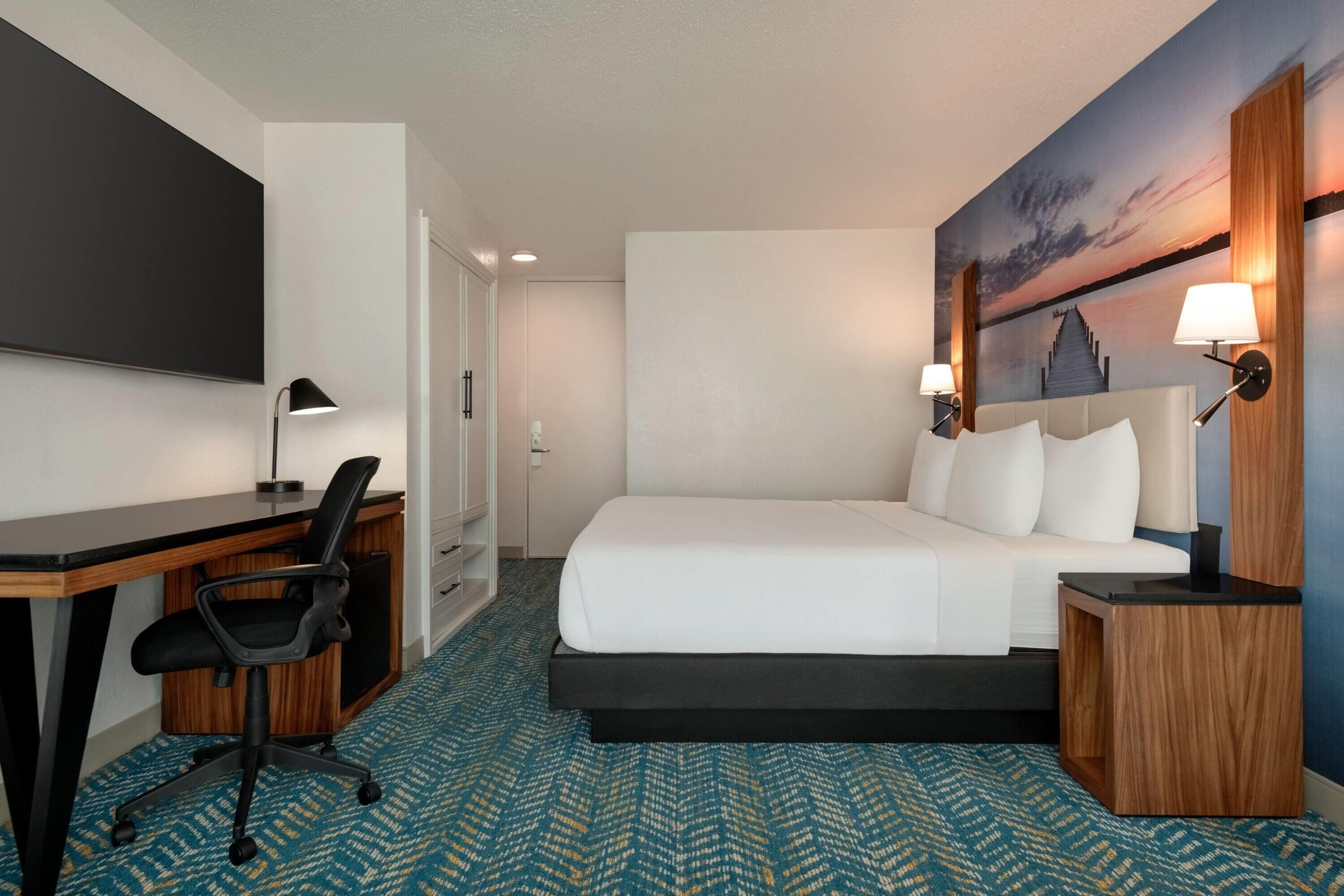 Quarto de hotel com cama, escrivaninha, cadeira e um quadro de pôr do sol na parede.