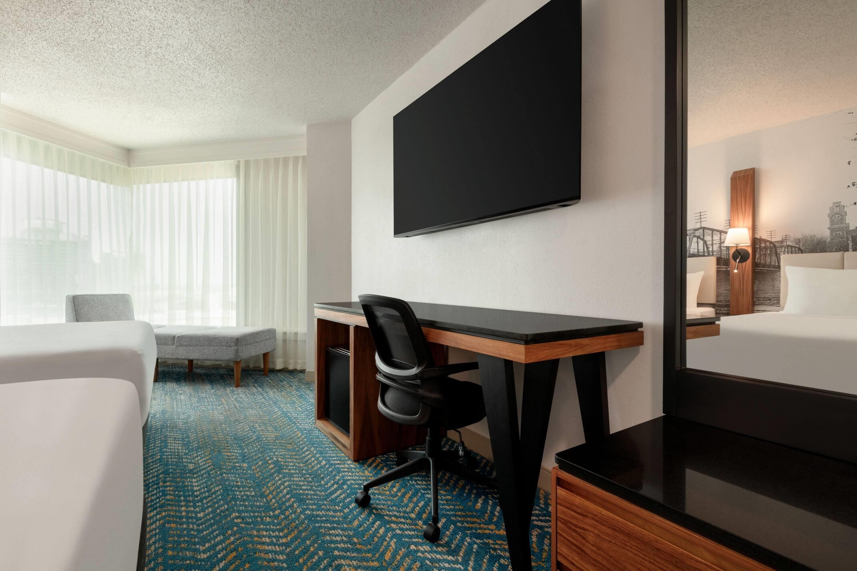 Quarto de hotel com uma cama grande, uma escrivaninha com cadeira, uma TV de tela plana e uma janela com cortinas translúcidas.