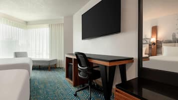 Quarto de hotel com uma cama grande, uma escrivaninha com cadeira, uma TV de tela plana e uma janela com cortinas translúcidas.