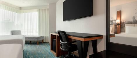 A hotel room with a large bed, a desk with a chair, a flat-screen TV, and a window with sheer curtains.