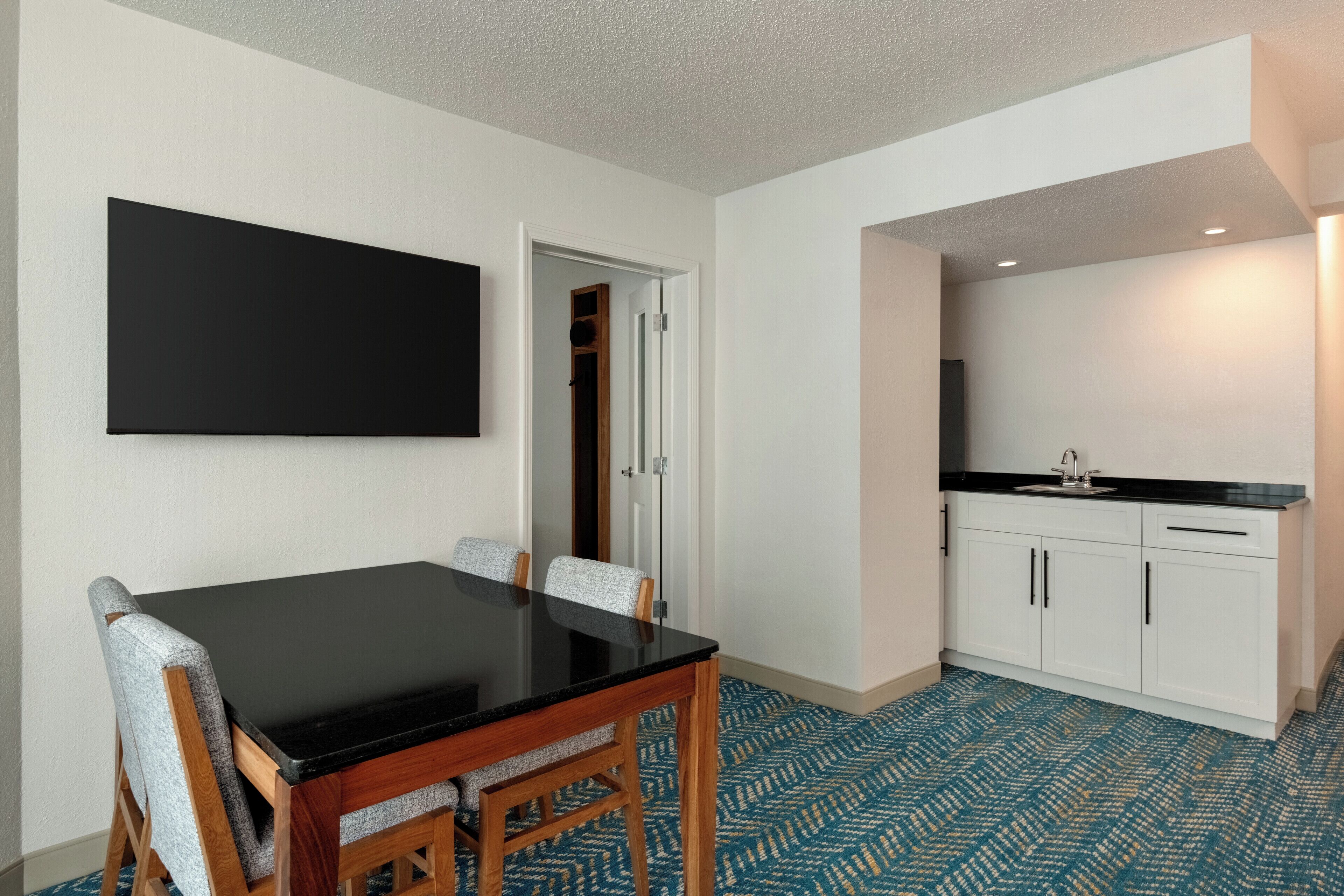 A compact room with a dining table, chairs, a kitchen area with cabinets, and a large black artwork on the wall.