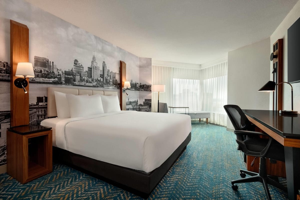 A hotel room with a large bed, a desk, a chair, and a cityscape mural on the wall.