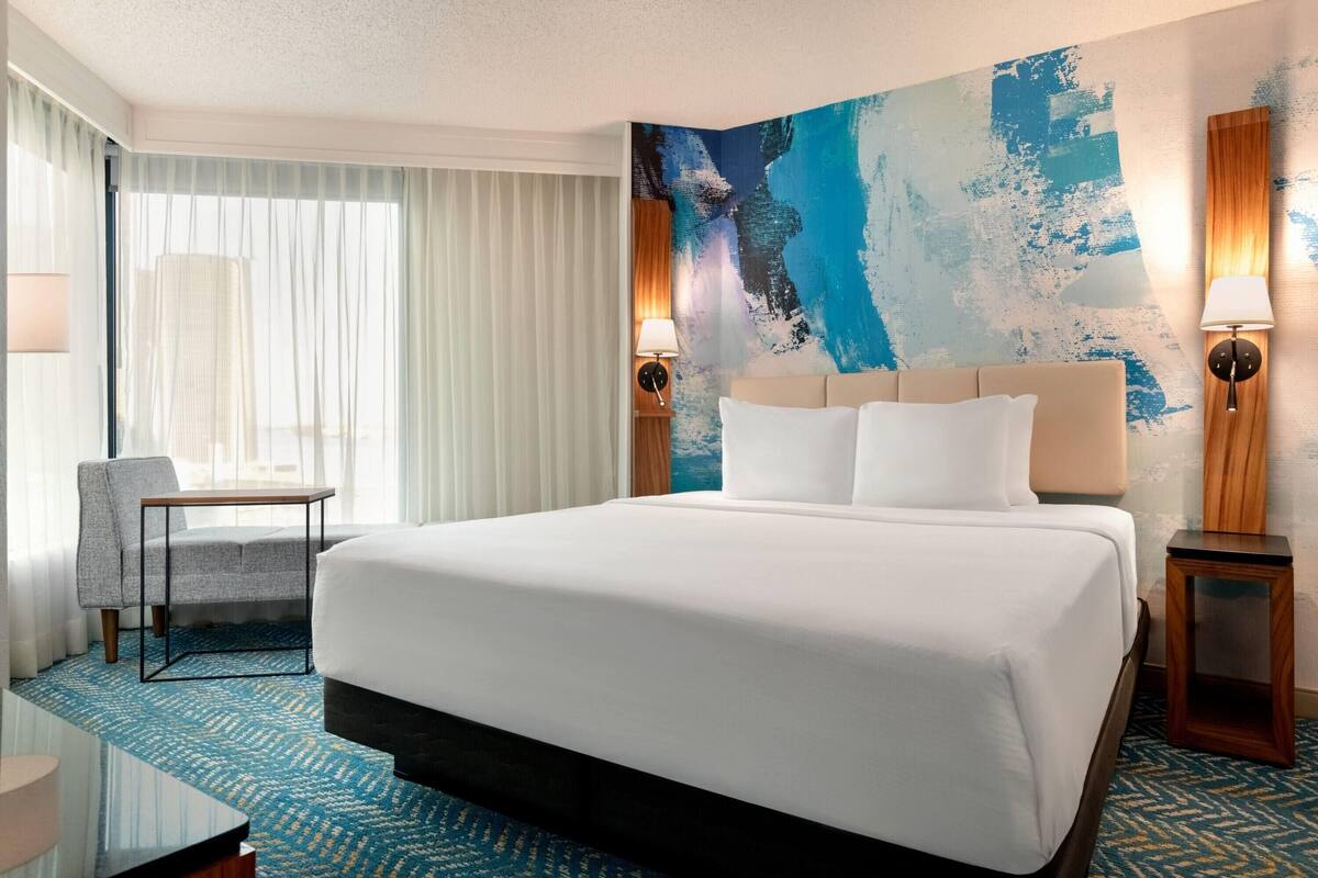 A hotel room with a large bed, a chair, a desk, and a wall with a blue abstract painting.