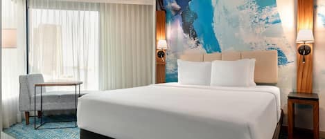 A hotel room with a large bed, a chair, a desk, and a wall with a blue abstract painting.