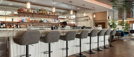 A modern bar area with a long counter, multiple stools, and a variety of bottles and glasses on shelves.