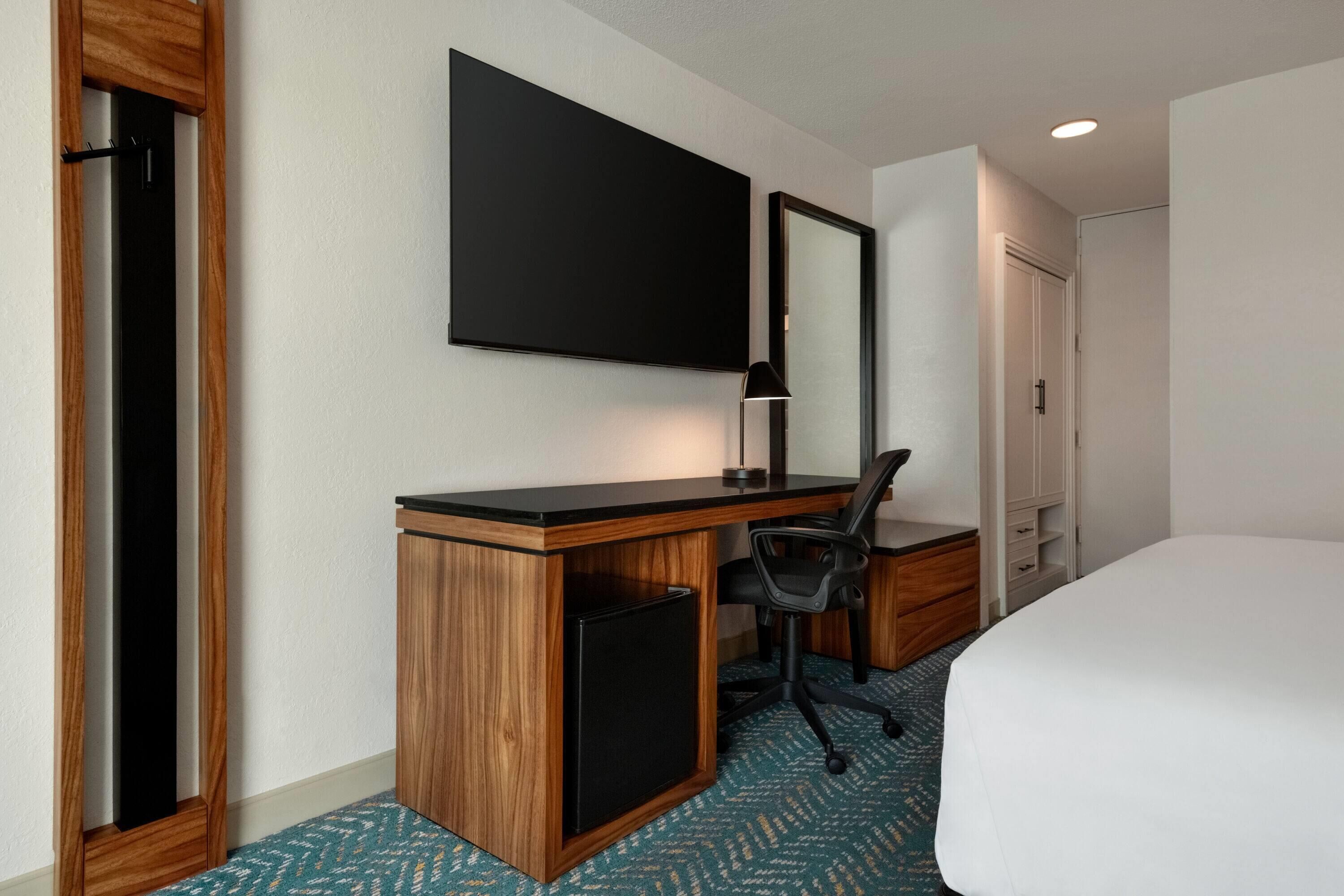 Une chambre d’hôtel avec un bureau en bois, une chaise noire, un lit, un grand miroir et un téléviseur à écran plat.