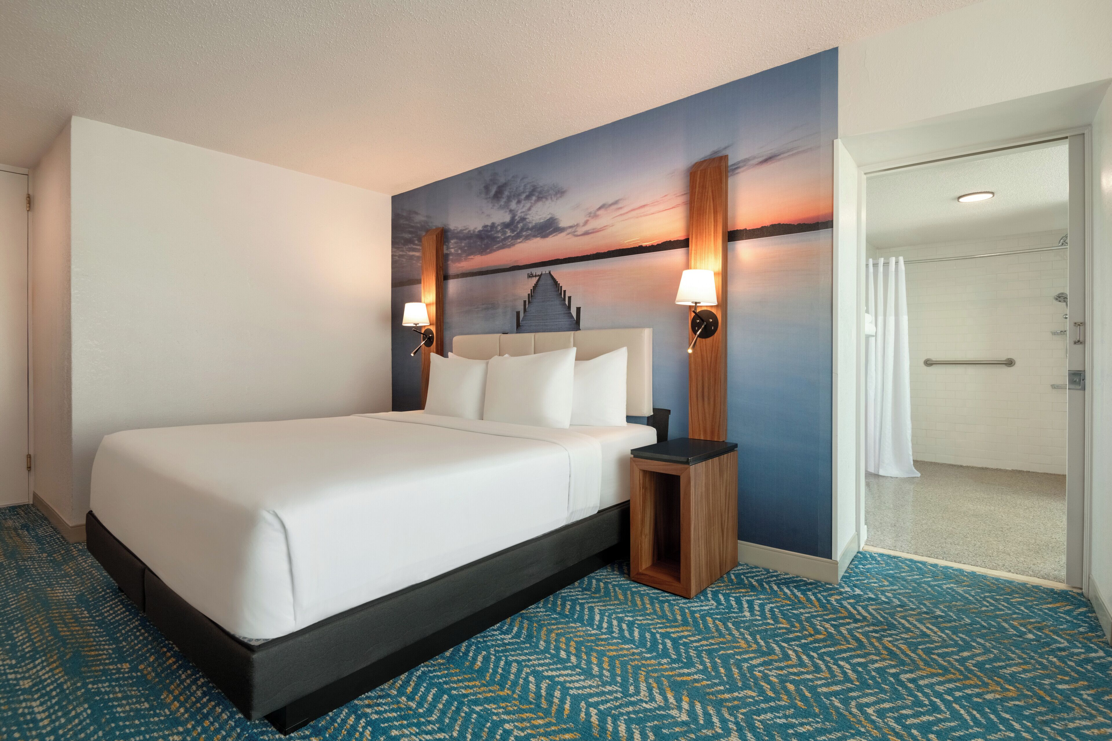 A hotel room with a large bed, a painting of a sunset on the wall, and a small wooden nightstand with two lamps.
