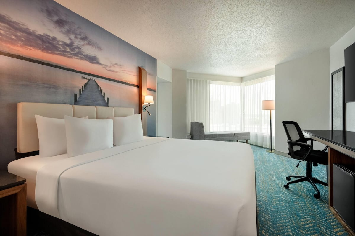 A hotel room with a large bed, a desk, a chair, and a view of a sunset over water.