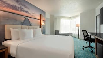 A hotel room with a large bed, a desk, a chair, and a view of a sunset over water.