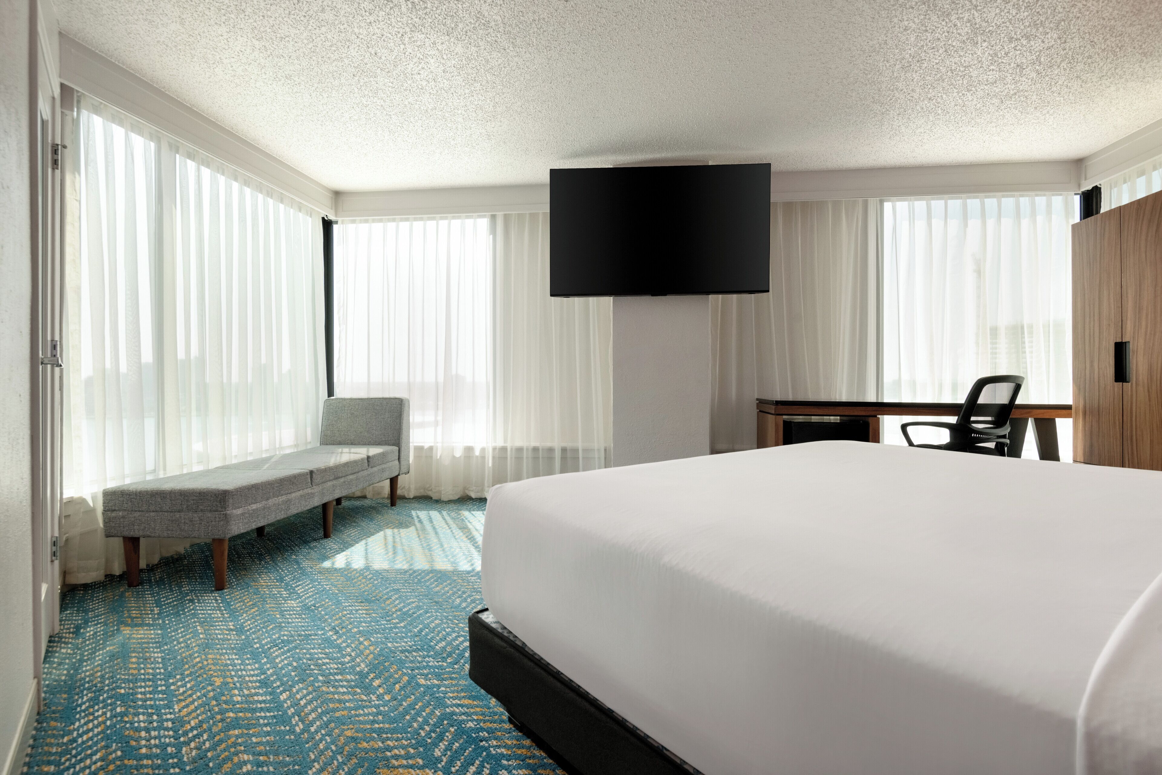 A hotel room with a large bed, a bench, a desk, a flat-screen TV, and large windows with sheer curtains.