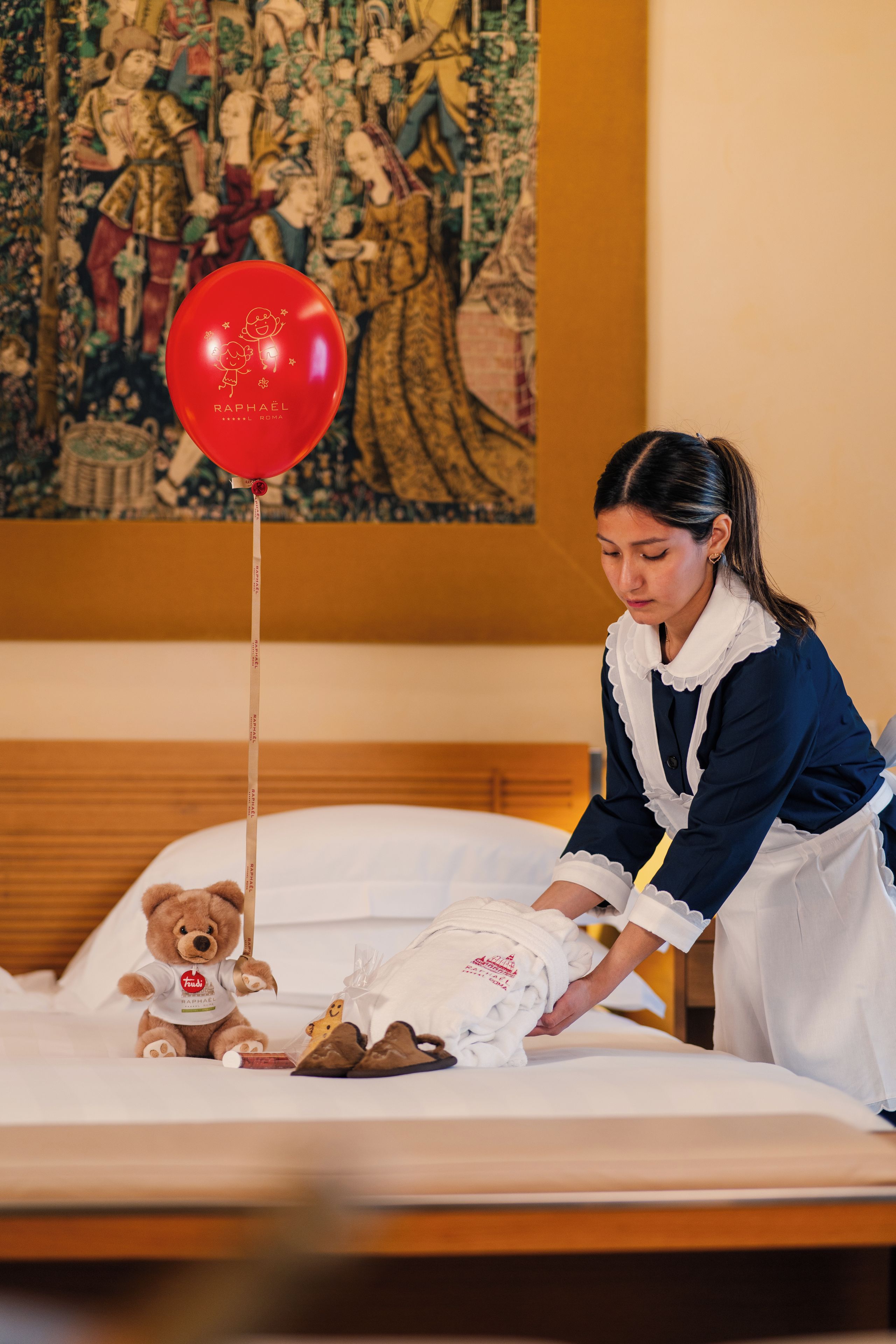 Ett hotellrum med en säng, en nallebjörn, en röd ballong och en tavla på väggen.
