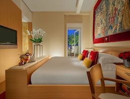 A modern hotel room with a large bed, a television, a small table with a plant, and a large tapestry on the wall.