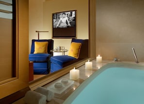 A modern bathroom with a bathtub, two blue chairs with yellow cushions, a small table with a teapot, and a framed black-and-white photo on the wall.