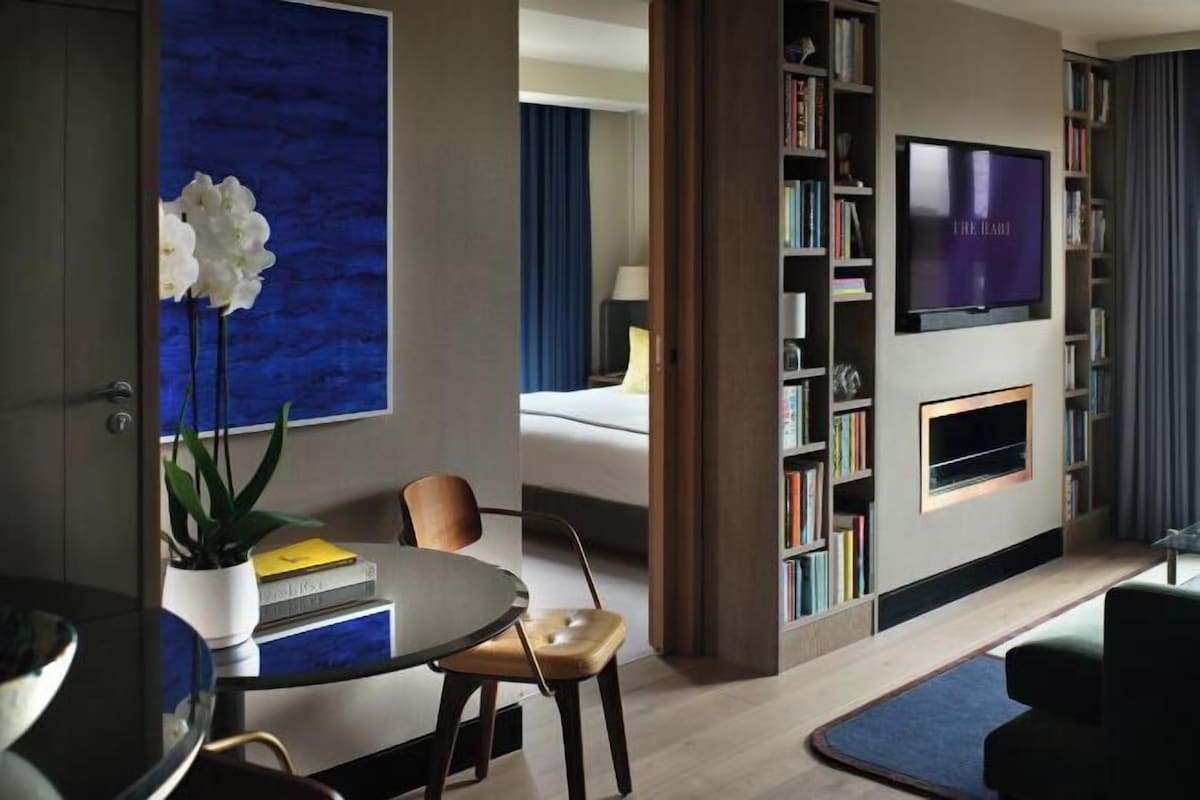 A modern hotel room with a dining area, a bookshelf, a flat-screen TV, and a large window.