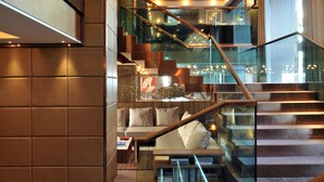 Un interior moderno con una escalera de cristal, barandillas de madera y una zona de estar con libros.