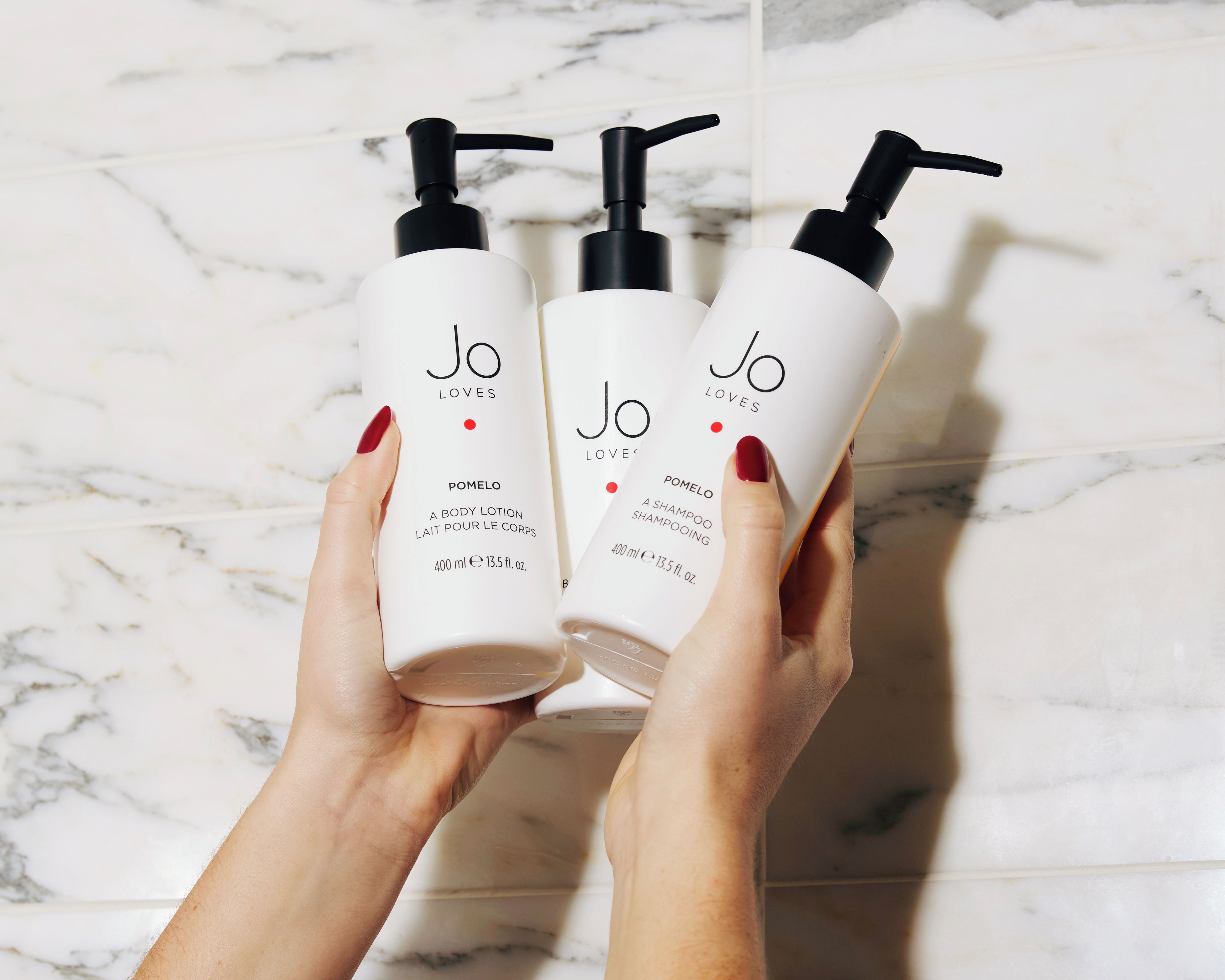 Trois flacons de lotion pour le corps et de shampoing de la marque Jo Loves, tenus dans des mains, sur un fond en marbre.