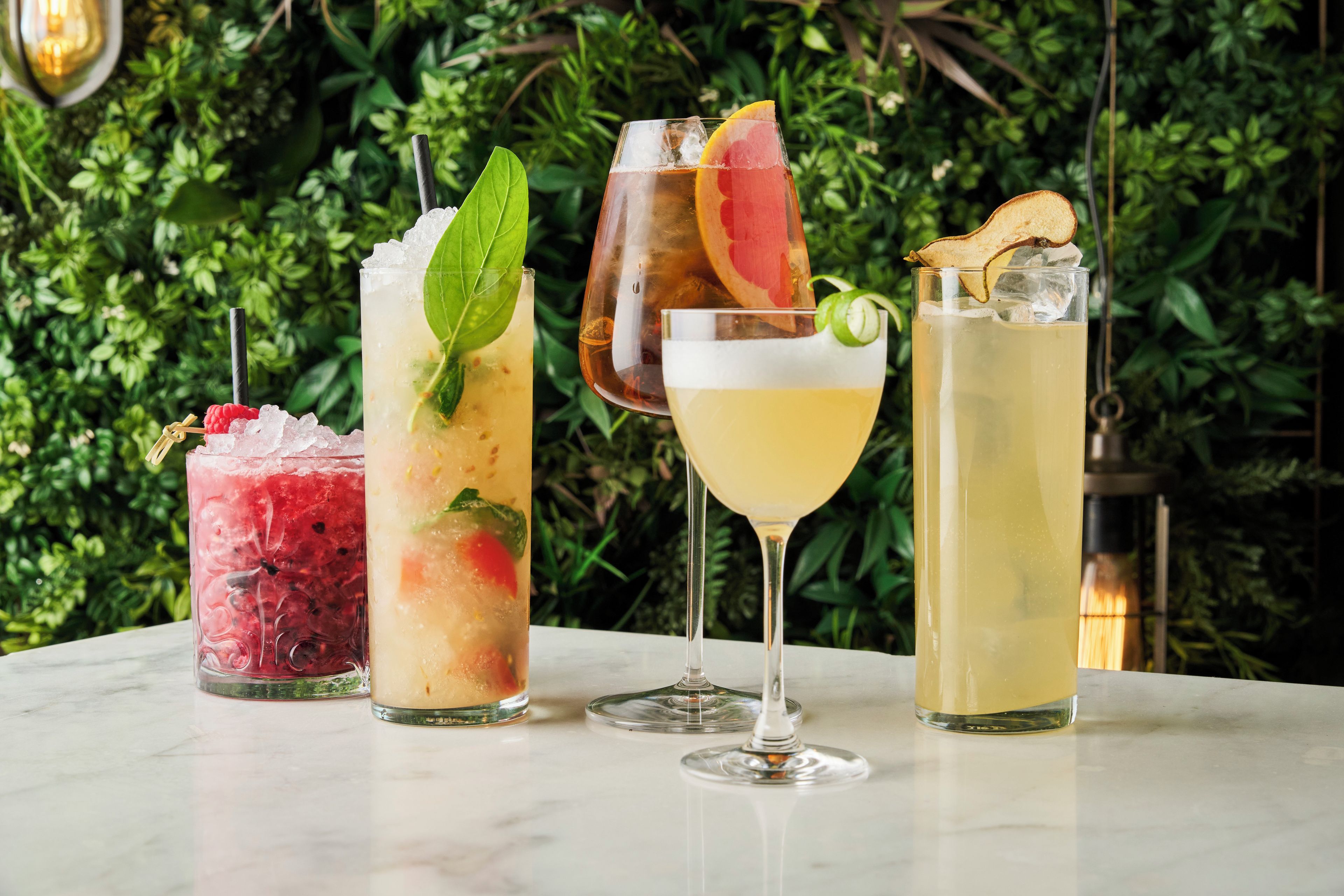Vier verschiedene Cocktails auf einem Marmortisch mit grüner Blattkulisse.