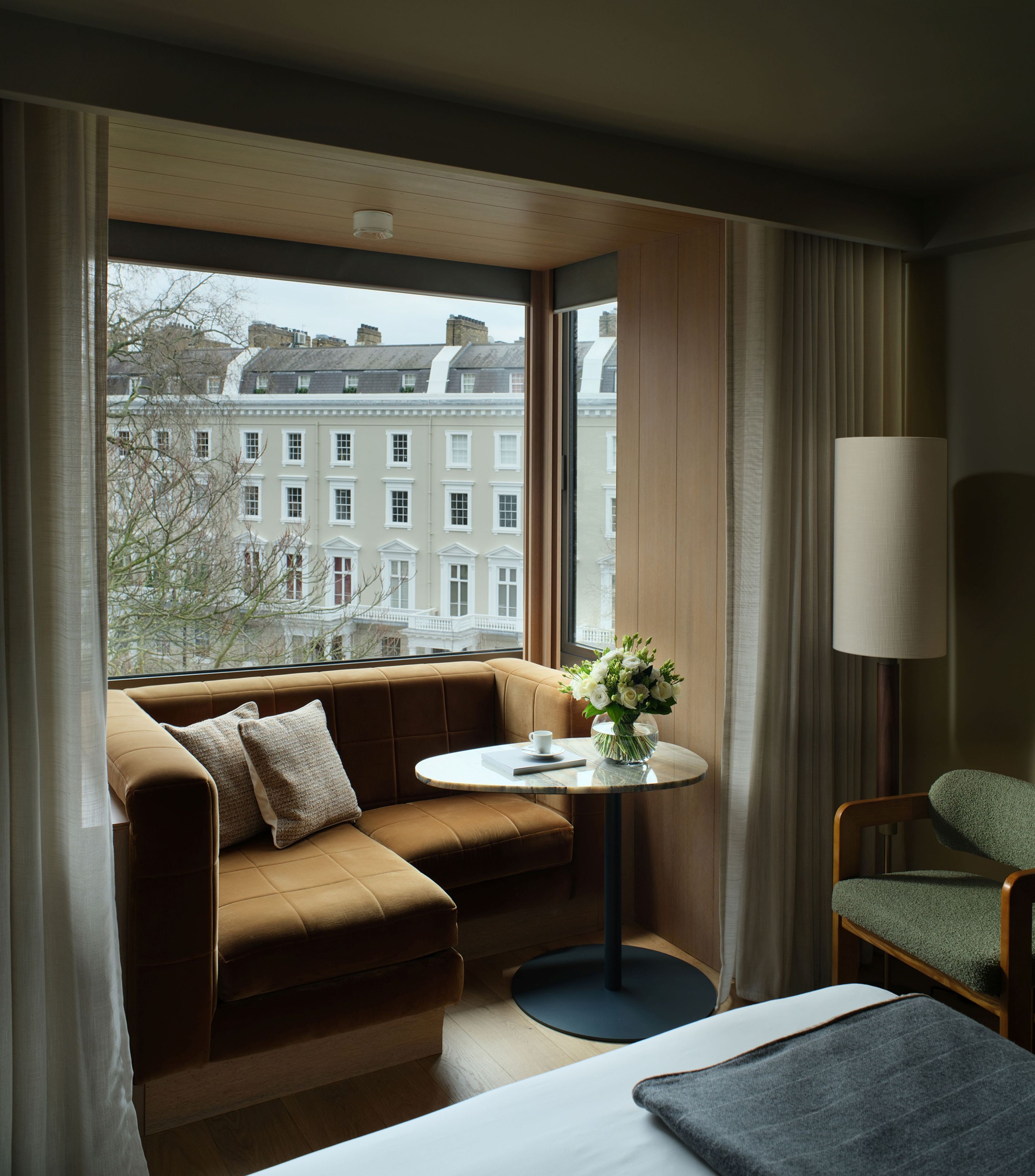 A hotel room with a sofa, a small table with a vase, a lamp, and a view of buildings outside.