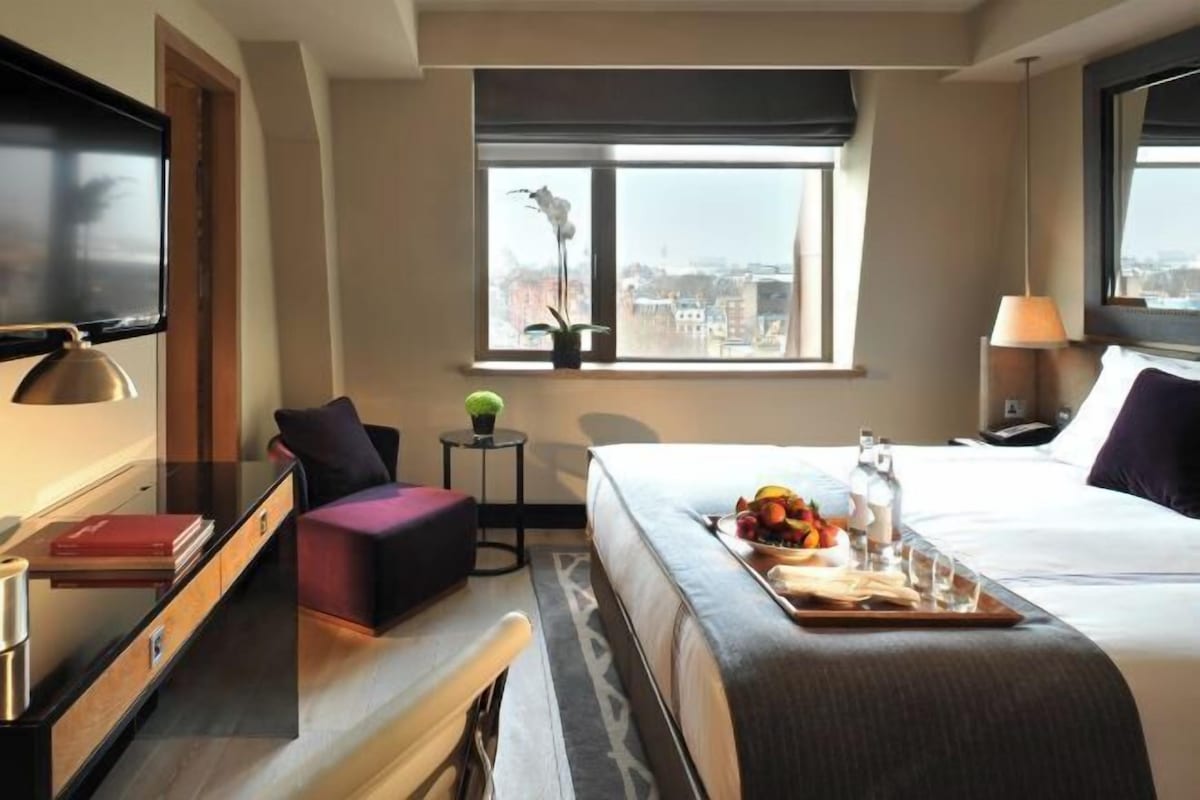 A hotel room with a bed, a desk, a chair, a small table with a tray of fruit, a TV, and a window with a city view.