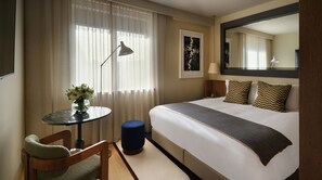 Habitación de hotel con cama, escritorio con lámpara, una silla y una mesita con un florero.